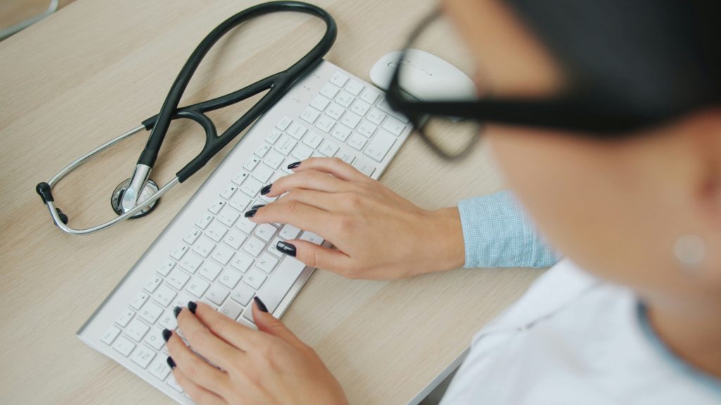 Healthcare professional typing at a desk with a stethoscope nearby